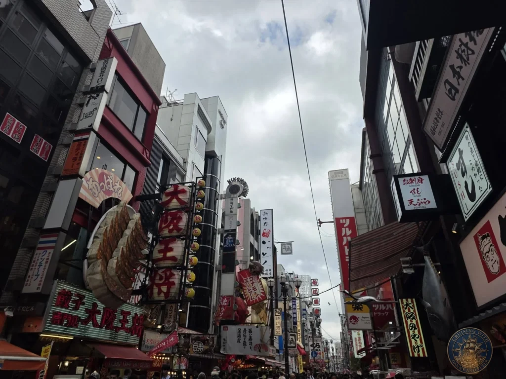 Kobe Japan Cruise Port Guide 2026 – Insider Tips & One Day Itinerary 43 Looking down the busy Dotonbori street in Osaka filled with colorful restaurant signs, neon lights, and crowds under a cloudy sky. Vibrant Osaka atmosphere from Kobe port day. Kobe Japan Cruise Port Guide | DIY Kobe Cruise Port to City Center | Things to do in Kobe from cruise ship | Westerdam Kobe Docking Location | One day in Kobe Japan itinerary by Danny and Peter.