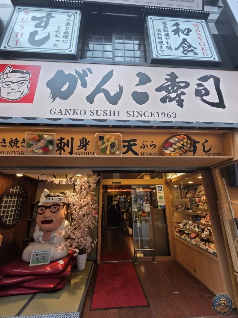 Kobe Japan Cruise Port Guide 2026 – Insider Tips & One Day Itinerary 5 Entrance to Ganko Sushi in Dotonbori featuring the large restaurant sign, mascot statue, and glass doors in Osaka, Japan. Popular sushi spot from Kobe port day. Kobe Japan Cruise Port Guide | DIY Kobe Cruise Port to City Center | Things to do in Kobe from cruise ship | Westerdam Kobe Docking Location | One day in Kobe Japan itinerary by Danny and Peter.