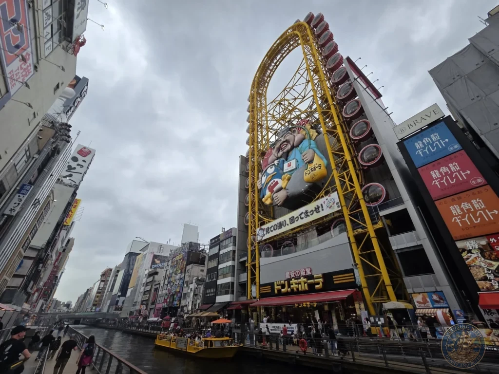 Kobe Japan Cruise Port Guide 2026 – Insider Tips & One Day Itinerary 99 Iconic Don Quijote store in Dotonbori featuring the large yellow Ferris wheel structure and giant character sign along the canal in Osaka, Japan. Famous shopping landmark from Kobe port day. Kobe Japan Cruise Port Guide | DIY Kobe Cruise Port to City Center | Things to do in Kobe from cruise ship | Westerdam Kobe Docking Location | One day in Kobe Japan itinerary by Danny and Peter.