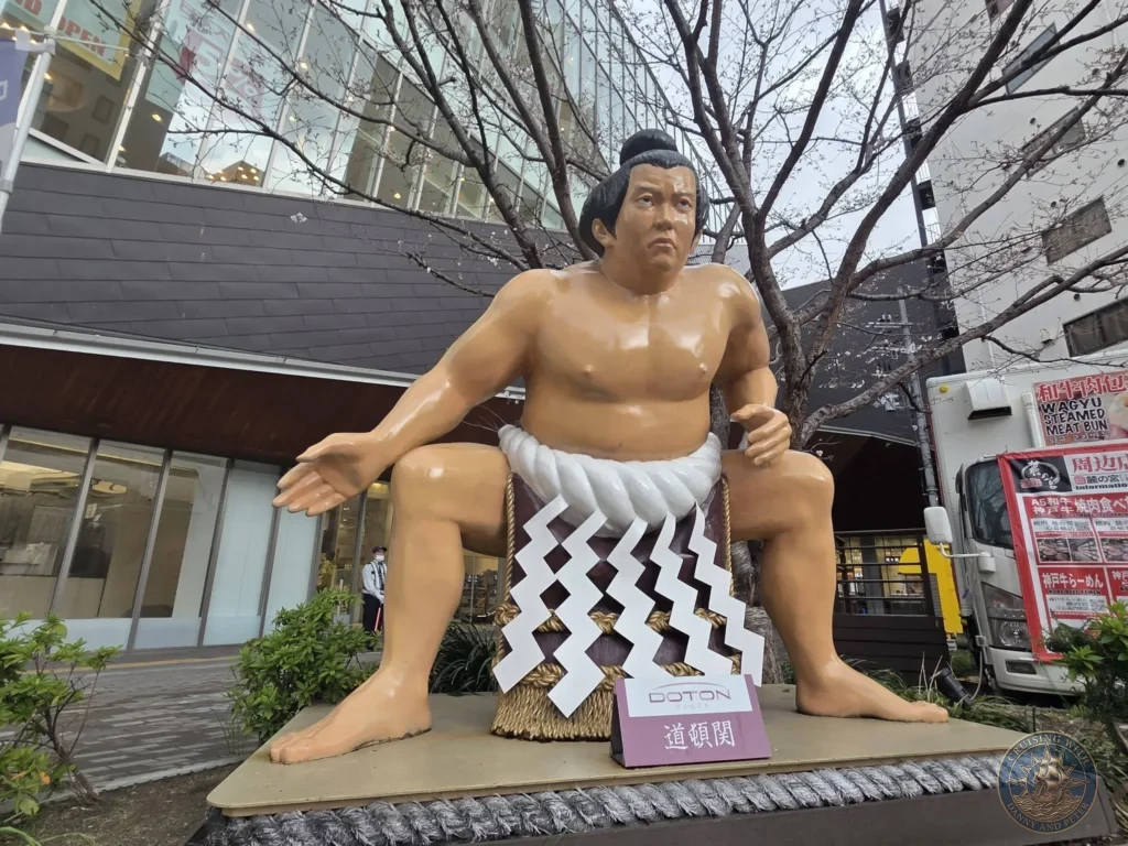Kobe Japan Cruise Port Guide 2026 – Insider Tips & One Day Itinerary 100 Large sumo wrestler statue seated on a platform at Doton Plaza in Dotonbori, Osaka, Japan. Fun landmark from Kobe port day. Kobe Japan Cruise Port Guide | DIY Kobe Cruise Port to City Center | Things to do in Kobe from cruise ship | Westerdam Kobe Docking Location | One day in Kobe Japan itinerary by Danny and Peter.