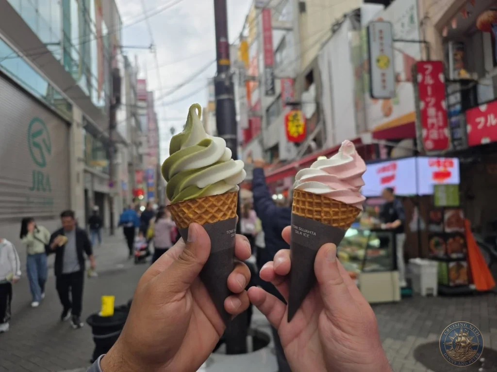 Kobe Japan Cruise Port Guide 2026 – Insider Tips & One Day Itinerary 101 Two soft serve ice cream cones, one matcha-vanilla swirl and one strawberry-vanilla, held up on a busy street in Osaka, Japan. Sweet treat from Kobe port day. Kobe Japan Cruise Port Guide | DIY Kobe Cruise Port to City Center | Things to do in Kobe from cruise ship | Westerdam Kobe Docking Location | One day in Kobe Japan itinerary by Danny and Peter.