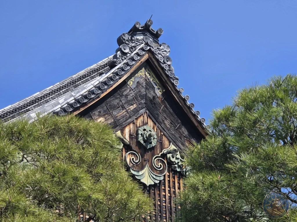 Kobe Japan Cruise Port Guide 2026 – Insider Tips & One Day Itinerary 150 Close-up of the ornate wooden gable and tiled roof of Nijo Castle with pine branches under a blue sky in Kyoto, Japan. Architectural detail from Kobe port day. Kobe Japan Cruise Port Guide | DIY Kobe Cruise Port to City Center | Things to do in Kobe from cruise ship | Westerdam Kobe Docking Location | One day in Kobe Japan itinerary by Danny and Peter.