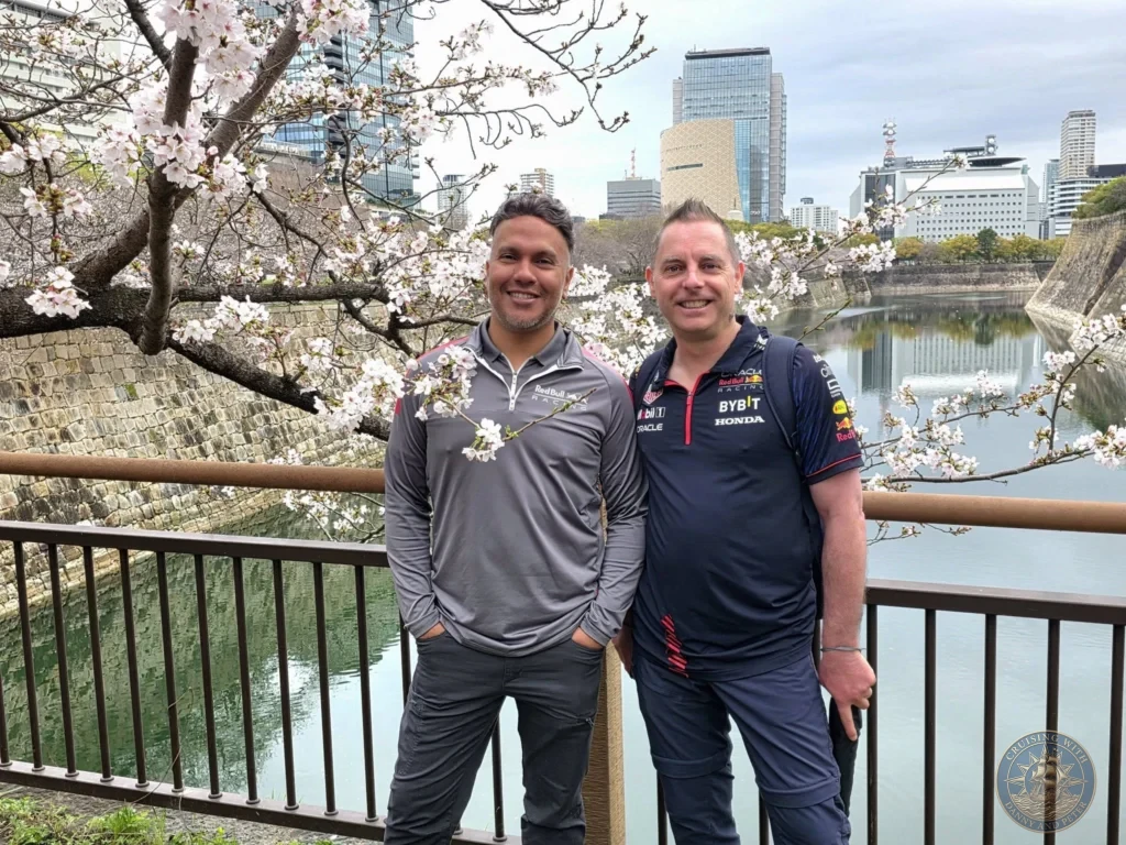 Kobe Japan Cruise Port Guide 2026 – Insider Tips & One Day Itinerary 102 Danny and Peter standing by the Osaka Castle moat with cherry blossoms blooming and the modern city skyline in the background in Osaka, Japan. Historic site from Kobe port day. Kobe Japan Cruise Port Guide | DIY Kobe Cruise Port to City Center | Things to do in Kobe from cruise ship | Westerdam Kobe Docking Location | One day in Kobe Japan itinerary by Danny and Peter.