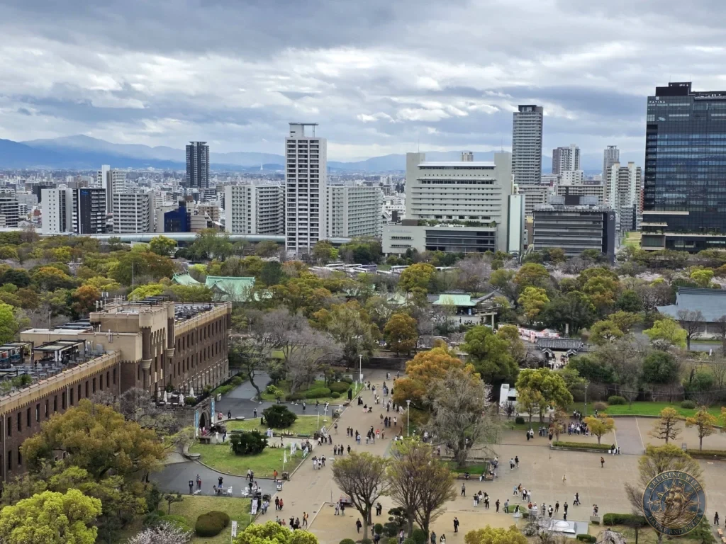 Kobe Japan Cruise Port Guide 2026 – Insider Tips & One Day Itinerary 109 Elevated view from Osaka Castle overlooking the busy park grounds, traditional buildings, trees and modern city skyline with mountains in the distance in Osaka, Japan. Panoramic scene from Kobe port day. Kobe Japan Cruise Port Guide | DIY Kobe Cruise Port to City Center | Things to do in Kobe from cruise ship | Westerdam Kobe Docking Location | One day in Kobe Japan itinerary by Danny and Peter.