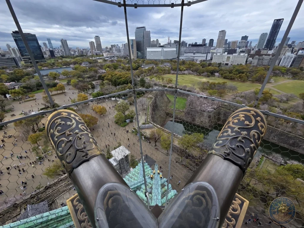 Kobe Japan Cruise Port Guide 2026 – Insider Tips & One Day Itinerary 110 View from the top of Osaka Castle looking past the golden shachihoko roof ornaments over the castle grounds, moat and modern city skyline in Osaka, Japan. Top-floor panorama from Kobe port day. Kobe Japan Cruise Port Guide | DIY Kobe Cruise Port to City Center | Things to do in Kobe from cruise ship | Westerdam Kobe Docking Location | One day in Kobe Japan itinerary by Danny and Peter.