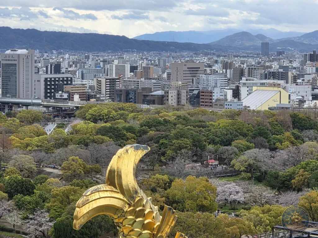 Kobe Japan Cruise Port Guide 2026 – Insider Tips & One Day Itinerary 111 View from Osaka Castle featuring a golden shachihoko ornament in the foreground with the park, city skyline and distant mountains beyond in Osaka, Japan. Iconic rooftop detail from Kobe port day. Kobe Japan Cruise Port Guide | DIY Kobe Cruise Port to City Center | Things to do in Kobe from cruise ship | Westerdam Kobe Docking Location | One day in Kobe Japan itinerary by Danny and Peter.