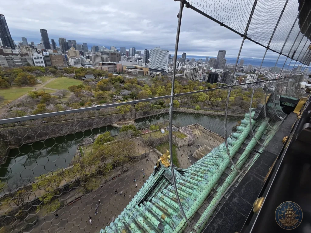 Kobe Japan Cruise Port Guide 2026 – Insider Tips & One Day Itinerary 113 View from the top of Osaka Castle looking down over the green-tiled roof, stone walls, moat, park grounds and modern city skyline in Osaka, Japan. High vantage point from Kobe port day. Kobe Japan Cruise Port Guide | DIY Kobe Cruise Port to City Center | Things to do in Kobe from cruise ship | Westerdam Kobe Docking Location | One day in Kobe Japan itinerary by Danny and Peter.