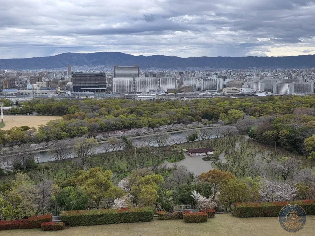 Kobe Japan Cruise Port Guide 2026 – Insider Tips & One Day Itinerary 114 Elevated view from Osaka Castle overlooking the park, moat, cherry blossoms and city skyline with mountains in the distance under cloudy skies in Osaka, Japan. Wide panorama from Kobe port day. Kobe Japan Cruise Port Guide | DIY Kobe Cruise Port to City Center | Things to do in Kobe from cruise ship | Westerdam Kobe Docking Location | One day in Kobe Japan itinerary by Danny and Peter.