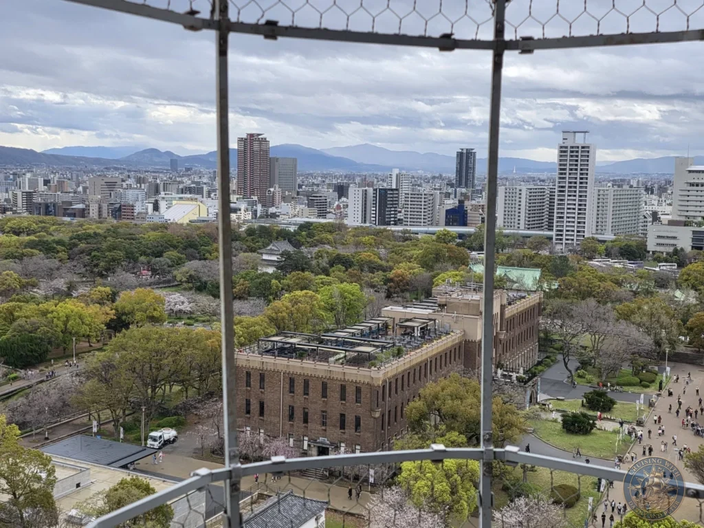 Kobe Japan Cruise Port Guide 2026 – Insider Tips & One Day Itinerary 115 View from Osaka Castle looking through the window mesh over the park, brick buildings, trees and city skyline with mountains in the distance in Osaka, Japan. Framed outlook from Kobe port day. Kobe Japan Cruise Port Guide | DIY Kobe Cruise Port to City Center | Things to do in Kobe from cruise ship | Westerdam Kobe Docking Location | One day in Kobe Japan itinerary by Danny and Peter.