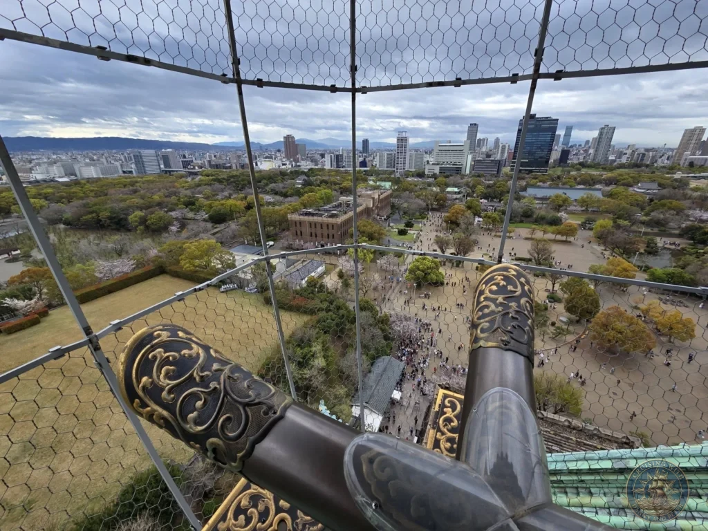 Kobe Japan Cruise Port Guide 2026 – Insider Tips & One Day Itinerary 116 View from the top of Osaka Castle looking past the golden roof ornaments down over the park grounds, crowds and modern city skyline in Osaka, Japan. High vantage point from Kobe port day. Kobe Japan Cruise Port Guide | DIY Kobe Cruise Port to City Center | Things to do in Kobe from cruise ship | Westerdam Kobe Docking Location | One day in Kobe Japan itinerary by Danny and Peter.