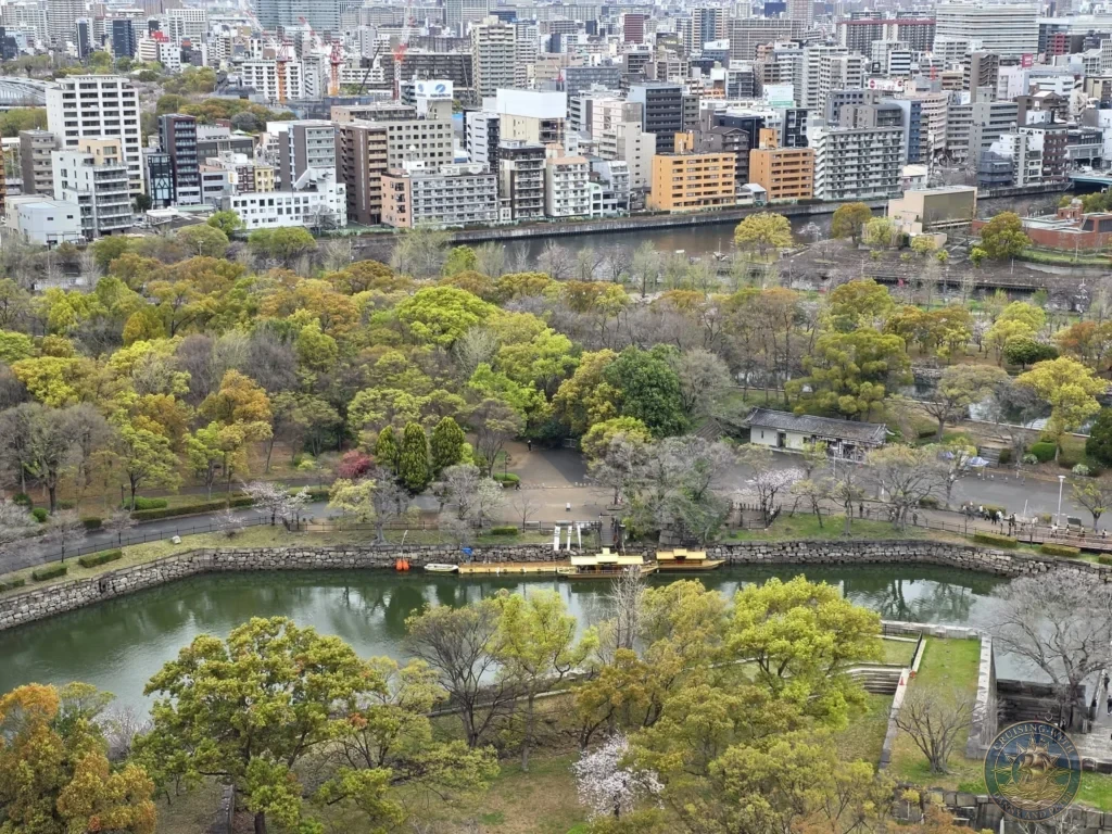 Kobe Japan Cruise Port Guide 2026 – Insider Tips & One Day Itinerary 117 Elevated view from Osaka Castle looking down over the moat with boats, park trees, stone walls and the dense city skyline beyond in Osaka, Japan. Scenic outlook from Kobe port day. Kobe Japan Cruise Port Guide | DIY Kobe Cruise Port to City Center | Things to do in Kobe from cruise ship | Westerdam Kobe Docking Location | One day in Kobe Japan itinerary by Danny and Peter.