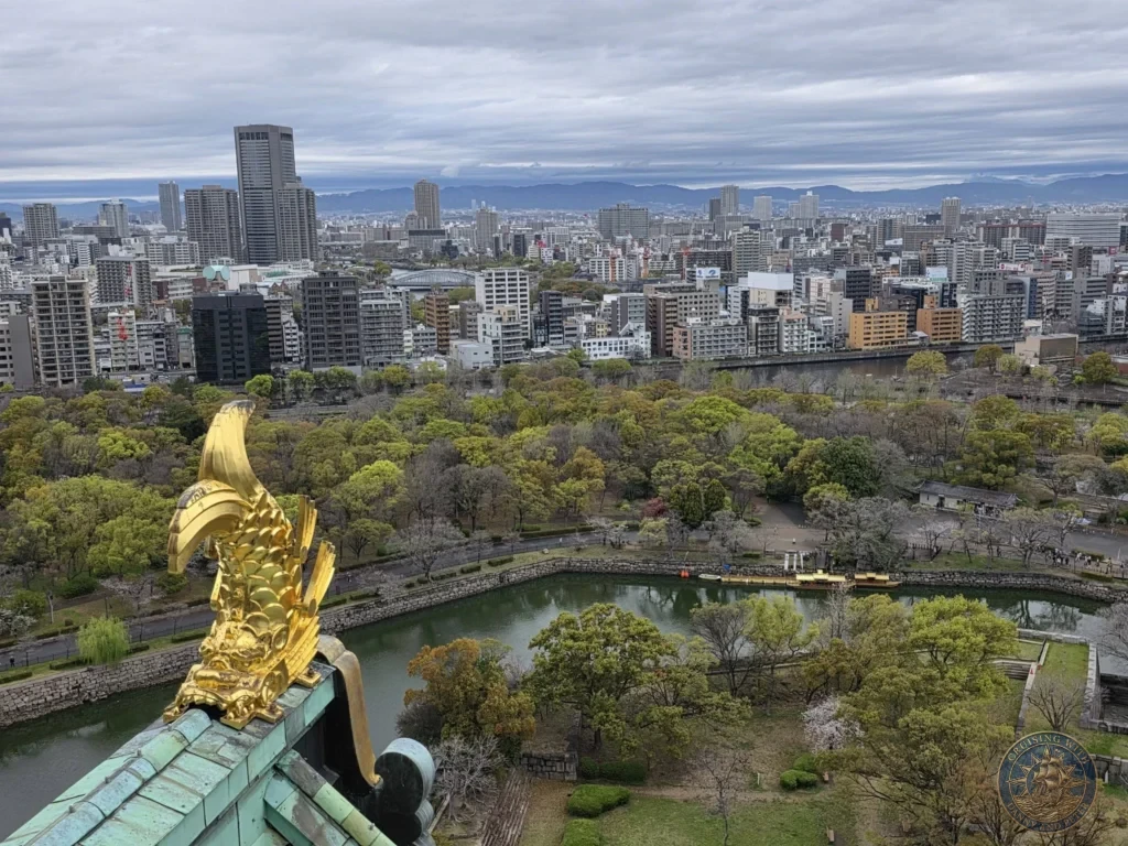 Kobe Japan Cruise Port Guide 2026 – Insider Tips & One Day Itinerary 118 View from the top of Osaka Castle featuring a golden shachihoko ornament in the foreground with the moat, park and expansive city skyline beyond in Osaka, Japan. Iconic rooftop detail from Kobe port day. Kobe Japan Cruise Port Guide | DIY Kobe Cruise Port to City Center | Things to do in Kobe from cruise ship | Westerdam Kobe Docking Location | One day in Kobe Japan itinerary by Danny and Peter.