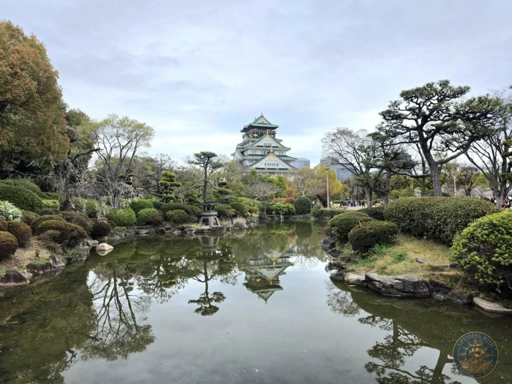 Kobe Japan Cruise Port Guide 2026 – Insider Tips & One Day Itinerary 11 Osaka Castle main keep reflected in a calm pond surrounded by manicured trees, bushes and a stone lantern in the castle gardens in Osaka, Japan. Classic garden view from Kobe port day. Kobe Japan Cruise Port Guide | DIY Kobe Cruise Port to City Center | Things to do in Kobe from cruise ship | Westerdam Kobe Docking Location | One day in Kobe Japan itinerary by Danny and Peter.