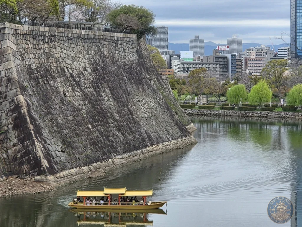 Kobe Japan Cruise Port Guide 2026 – Insider Tips & One Day Itinerary 125 A traditional yellow boat carrying passengers cruising along the wide water moat below the massive stone ramparts of Osaka Castle, with the modern city skyline in the background on an overcast spring day; ideal for a Kobe Japan Cruise Port Guide, DIY Kobe Cruise Port to City Center, Things to do in Kobe from cruise ship, Westerdam Kobe Docking Location, or One day in Kobe Japan itinerary, by Danny and Peter.