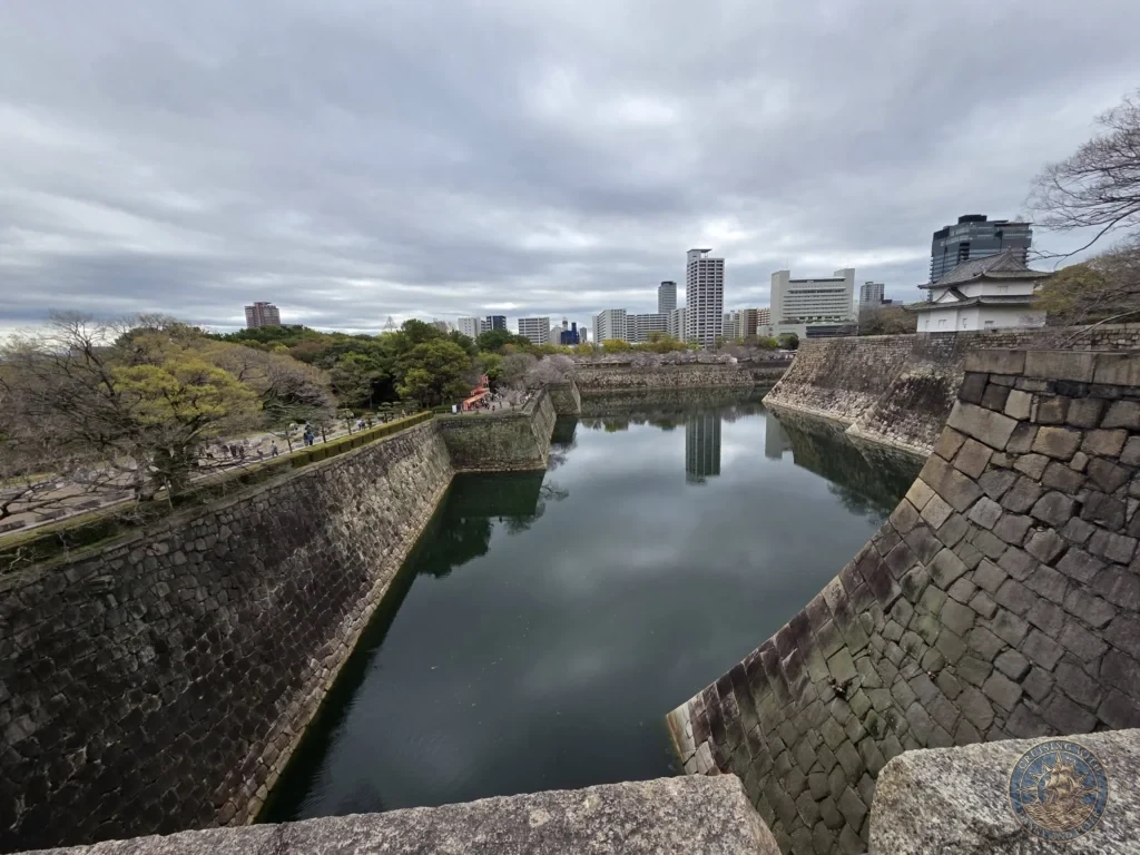 Kobe Japan Cruise Port Guide 2026 – Insider Tips & One Day Itinerary 126 A wide view of the massive stone walls surrounding the outer moat of Osaka Castle, reflecting modern city skyscrapers and overcast skies in the water below; ideal for a Kobe Japan Cruise Port Guide, DIY Kobe Cruise Port to City Center, Things to do in Kobe from cruise ship, Westerdam Kobe Docking Location, or One day in Kobe Japan itinerary, by Danny and Peter.