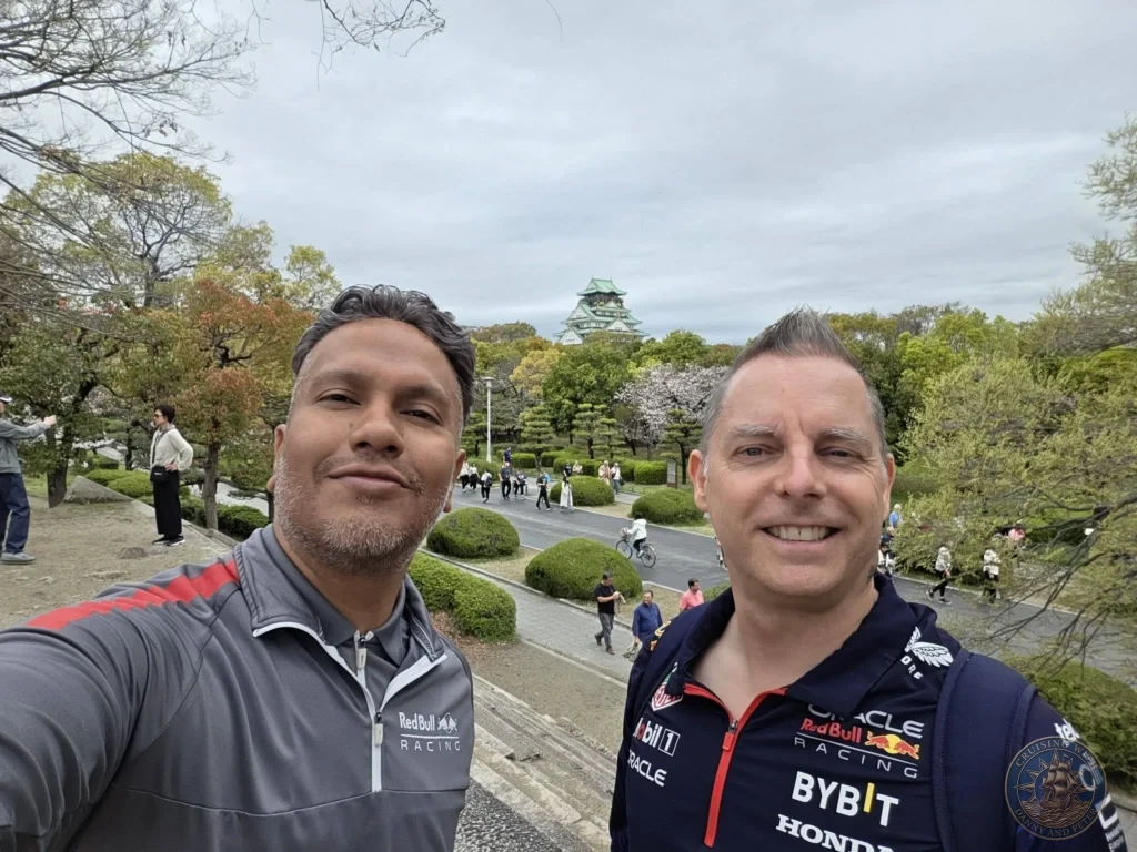 Kobe Japan Cruise Port Guide 2026 – Insider Tips & One Day Itinerary 127 Danny and Peter, wearing Red Bull Racing gear (a gray zip-up and a dark blue Oracle Bybit Honda polo), smile for a selfie on stone steps with Osaka Castle in the background under an overcast spring sky; ideal for a Kobe Japan Cruise Port Guide, DIY Kobe Cruise Port to City Center, Things to do in Kobe from cruise ship, Westerdam Kobe Docking Location, or One day in Kobe Japan itinerary, by Danny and Peter.