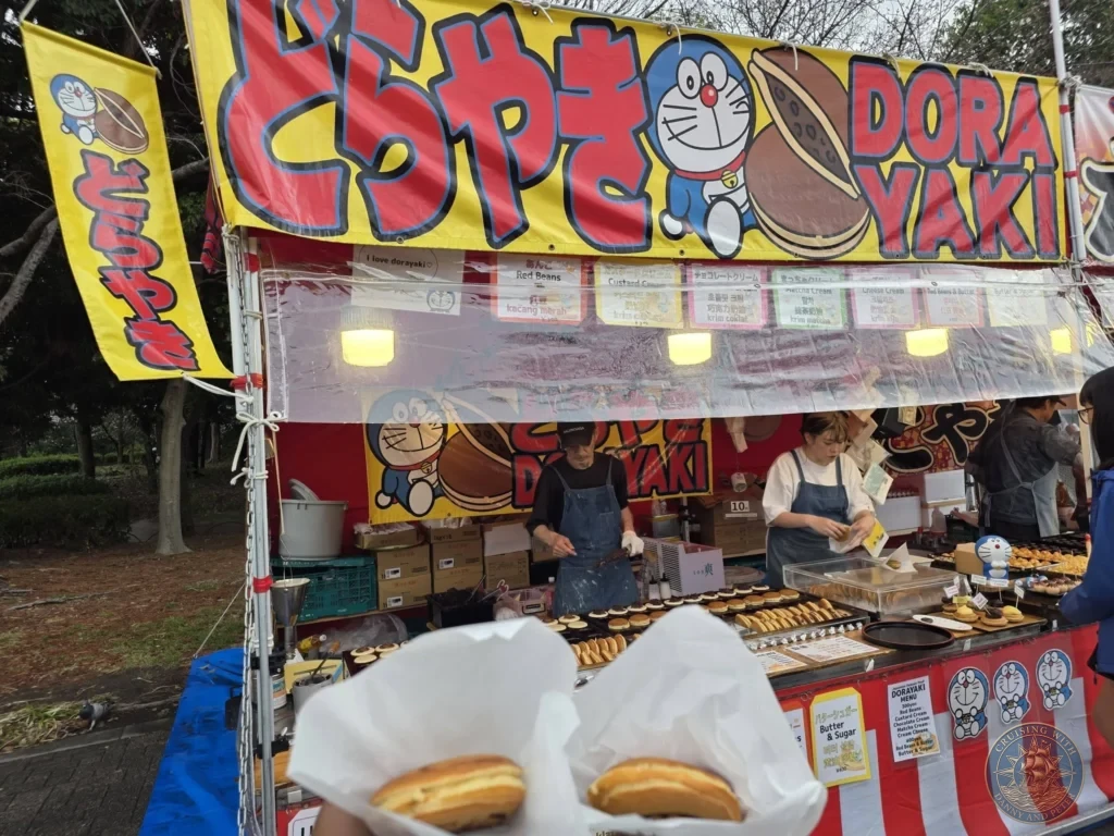 Kobe Japan Cruise Port Guide 2026 – Insider Tips & One Day Itinerary 128 Two warm dorayaki pancakes held in paper wrappers in the foreground, with a colorful Doraemon-themed street food stall and vendors freshly baking Japanese sweet red bean treats in Osaka Castle Park; ideal for a Kobe Japan Cruise Port Guide, DIY Kobe Cruise Port to City Center, Things to do in Kobe from cruise ship, Westerdam Kobe Docking Location, or One day in Kobe Japan itinerary, by Danny and Peter.