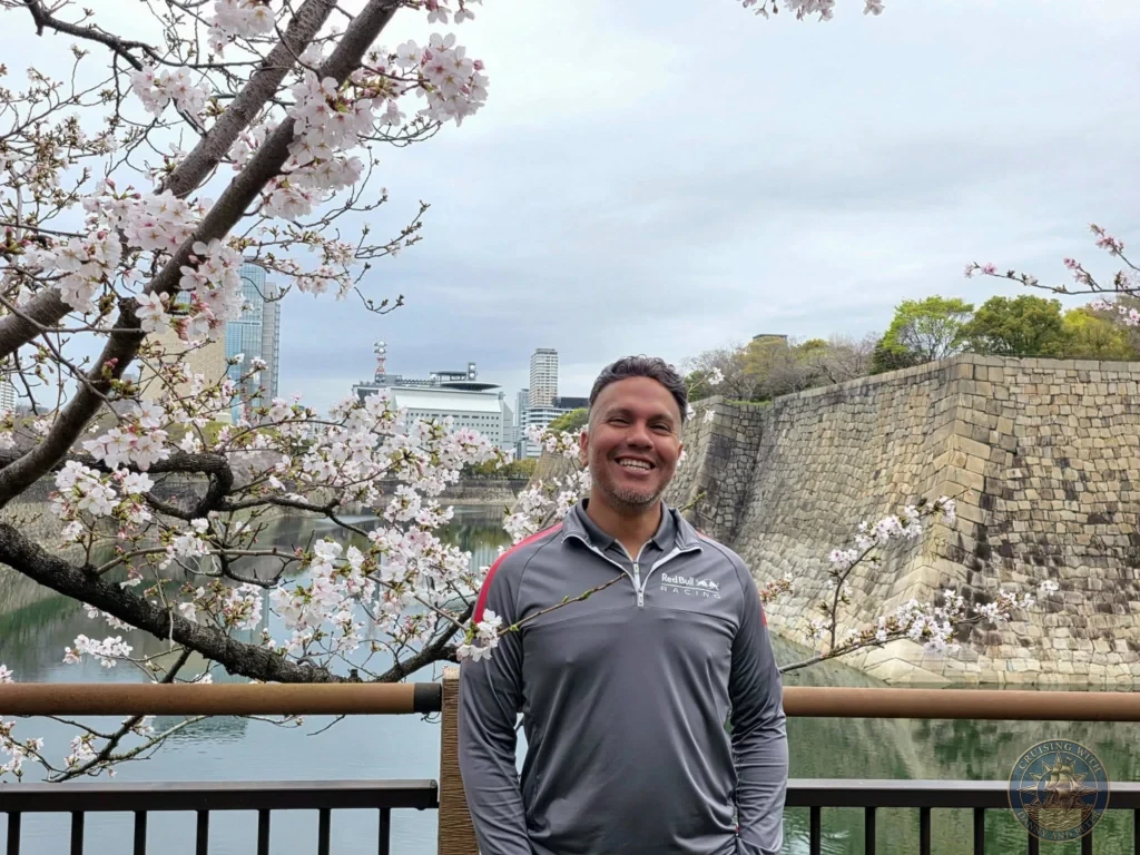 Kobe Japan Cruise Port Guide 2026 – Insider Tips & One Day Itinerary 130 Danny smiling in front of a railings overlook with blooming cherry blossoms framing the Osaka Castle moat, stone ramparts, and distant city skyline on a spring day; ideal for a Kobe Japan Cruise Port Guide, DIY Kobe Cruise Port to City Center, Things to do in Kobe from cruise ship, Westerdam Kobe Docking Location, or One day in Kobe Japan itinerary, by Danny and Peter.