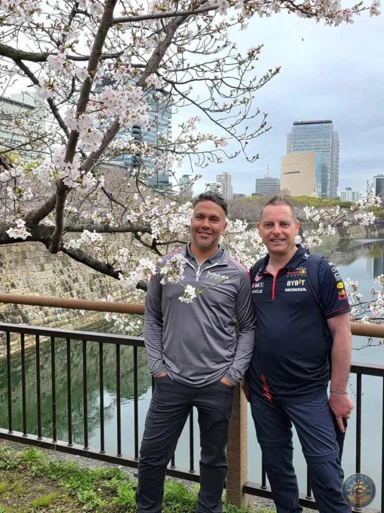 Kobe Japan Cruise Port Guide 2026 – Insider Tips & One Day Itinerary 131 Danny and Peter in Red Bull Racing shirts smiling together by a railing, framed by blooming cherry blossoms over the Osaka Castle moat with modern city skyscrapers across the water; ideal for a Kobe Japan Cruise Port Guide, DIY Kobe Cruise Port to City Center, Things to do in Kobe from cruise ship, Westerdam Kobe Docking Location, or One day in Kobe Japan itinerary, by Danny and Peter.