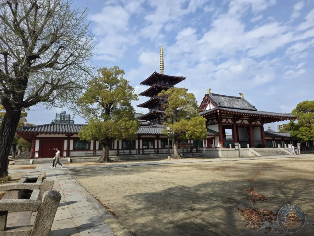 Kobe Japan Cruise Port Guide 2026 – Insider Tips & One Day Itinerary 45 A wide view of the five-story pagoda and traditional red-and-white wooden temple halls of Shitennoji Temple surrounded by green trees under a bright blue sky with light clouds; ideal for a Kobe Japan Cruise Port Guide, DIY Kobe Cruise Port to City Center, Things to do in Kobe from cruise ship, Westerdam Kobe Docking Location, or One day in Kobe Japan itinerary, by Danny and Peter.