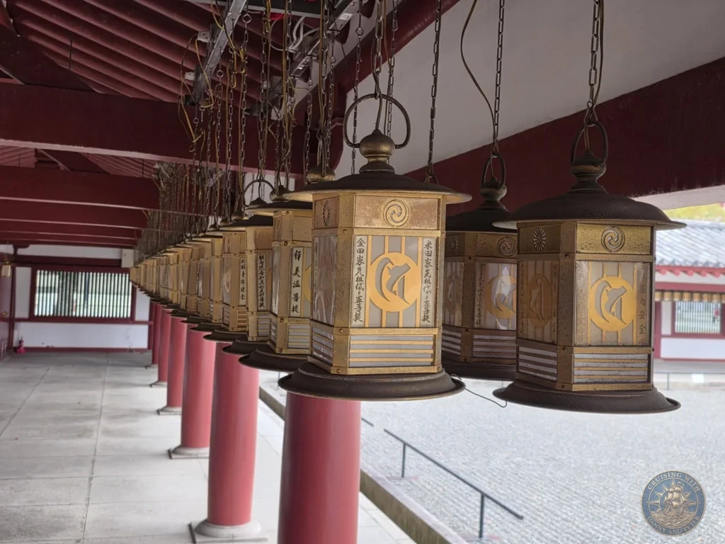 Kobe Japan Cruise Port Guide 2026 – Insider Tips & One Day Itinerary 136 A row of ornate golden metal hanging lanterns with Japanese calligraphy suspended along a covered red corridor at Shitennoji Temple in Osaka, Japan; ideal for a Kobe Japan Cruise Port Guide, DIY Kobe Cruise Port to City Center, Things to do in Kobe from cruise ship, MS Westerdam Kobe Docking Location, or One day in Kobe Japan itinerary, by Danny and Peter.