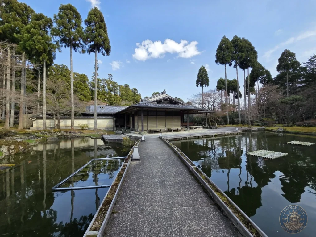 Kobe Japan Cruise Port Guide 2026 – Insider Tips & One Day Itinerary 33 Pathway leading across a calm pond toward a traditional Japanese building surrounded by tall trees under a blue sky at Shozan Resort Garden in Kyoto, Japan. Peaceful garden view from Kobe port day. Kobe Japan Cruise Port Guide | DIY Kobe Cruise Port to City Center | Things to do in Kobe from cruise ship | Westerdam Kobe Docking Location | One day in Kobe Japan itinerary by Danny and Peter.
