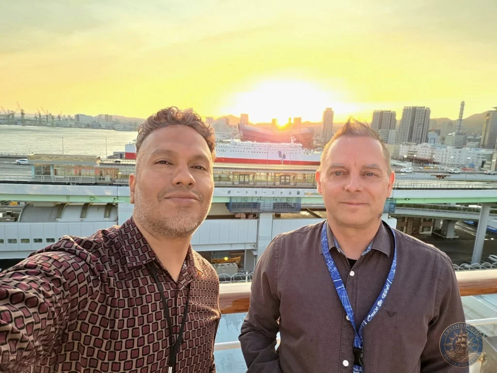 Kobe Japan Cruise Port Guide 2026 – Insider Tips & One Day Itinerary 19 Danny taking a selfie with Peter standing beside him on the deck of the cruise ship, with a vibrant glowing sunset over Kobe harbor and Port Terminal Station in the background; ideal for a Kobe Japan Cruise Port Guide, DIY Kobe Cruise Port to City Center, Things to do in Kobe from cruise ship, MS Westerdam Kobe Docking Location, or One day in Kobe Japan itinerary, by Danny and Peter.