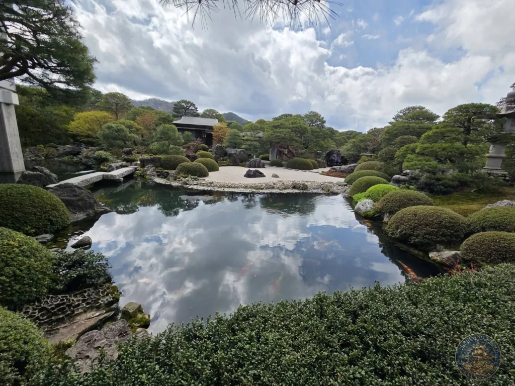 Tranquil Japanese koi pond reflecting dramatic blue sky and white clouds, framed by stone bridges, sculpted bushes, and raked sand at the Adachi Museum of Art during a shore excursion from Sakaiminato Japan Cruise Port Guide, DIY Sakaiminato Cruise Port to City Center, Things to do in Sakaiminato from cruise ship, Westerdam Sakaiminato Docking Location, One day in Sakaiminato Japan itinerary by Danny and Peter.