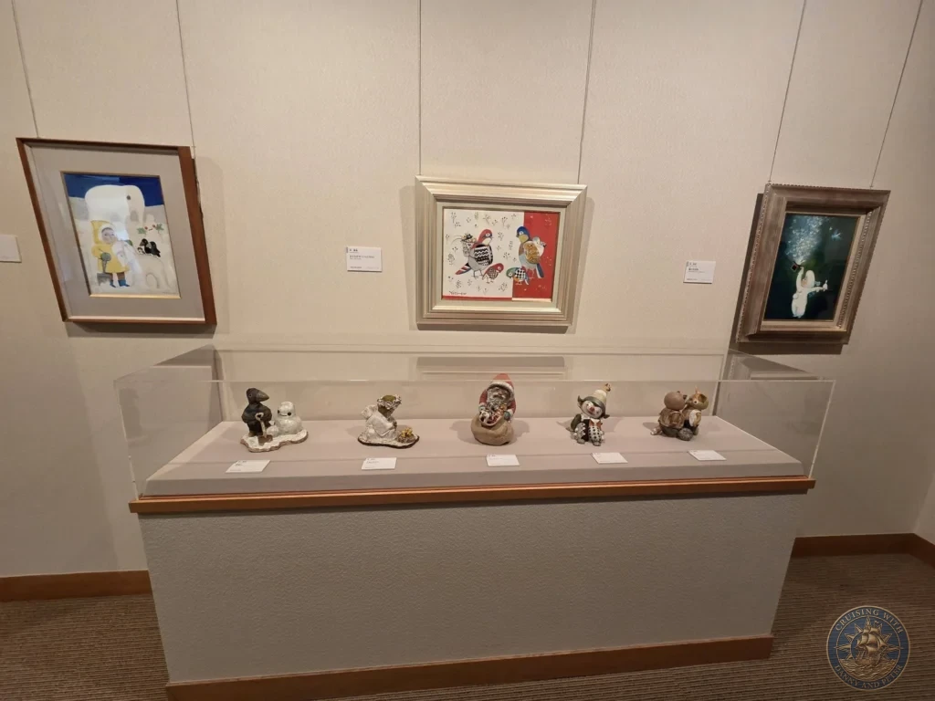 Framed paintings hanging on museum wall above a glass showcase displaying small handcrafted ceramic figurines at the Adachi Museum of Art during a shore excursion from Sakaiminato Japan Cruise Port Guide, DIY Sakaiminato Cruise Port to City Center, Things to do in Sakaiminato from cruise ship, Westerdam Sakaiminato Docking Location, One day in Sakaiminato Japan itinerary by Danny and Peter.