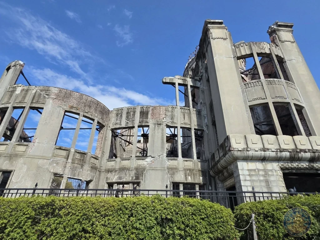 Hiroshima Japan Cruise Port Guide 2026 – Insider Tips & One Day Itinerary 12 Detailed view of the ruined concrete exterior and exposed brickwork of the Atomic Bomb Dome behind a green hedge under a clear blue sky in Hiroshima. Hiroshima Japan Cruise Port Guide, DIY Hiroshima Cruise Port to City Center, Things to do in Hiroshima from cruise ship, Westerdam Hiroshima Docking Location, One day in Hiroshima Japan itinerary by Danny and Peter.