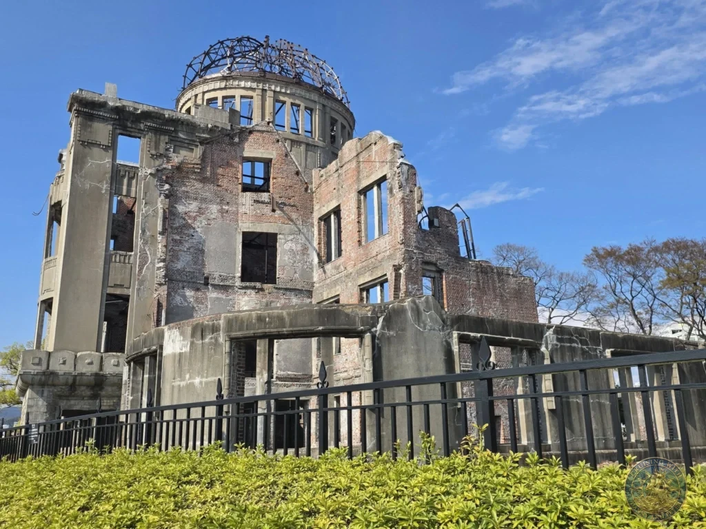Hiroshima Japan Cruise Port Guide 2026 – Insider Tips & One Day Itinerary 16 Angled shot of the Atomic Bomb Dome showing exposed red brick walls and steel skeleton dome behind a black fence and green hedge in Hiroshima. Hiroshima Japan Cruise Port Guide, DIY Hiroshima Cruise Port to City Center, Things to do in Hiroshima from cruise ship, Westerdam Hiroshima Docking Location, One day in Hiroshima Japan itinerary by Danny and Peter.