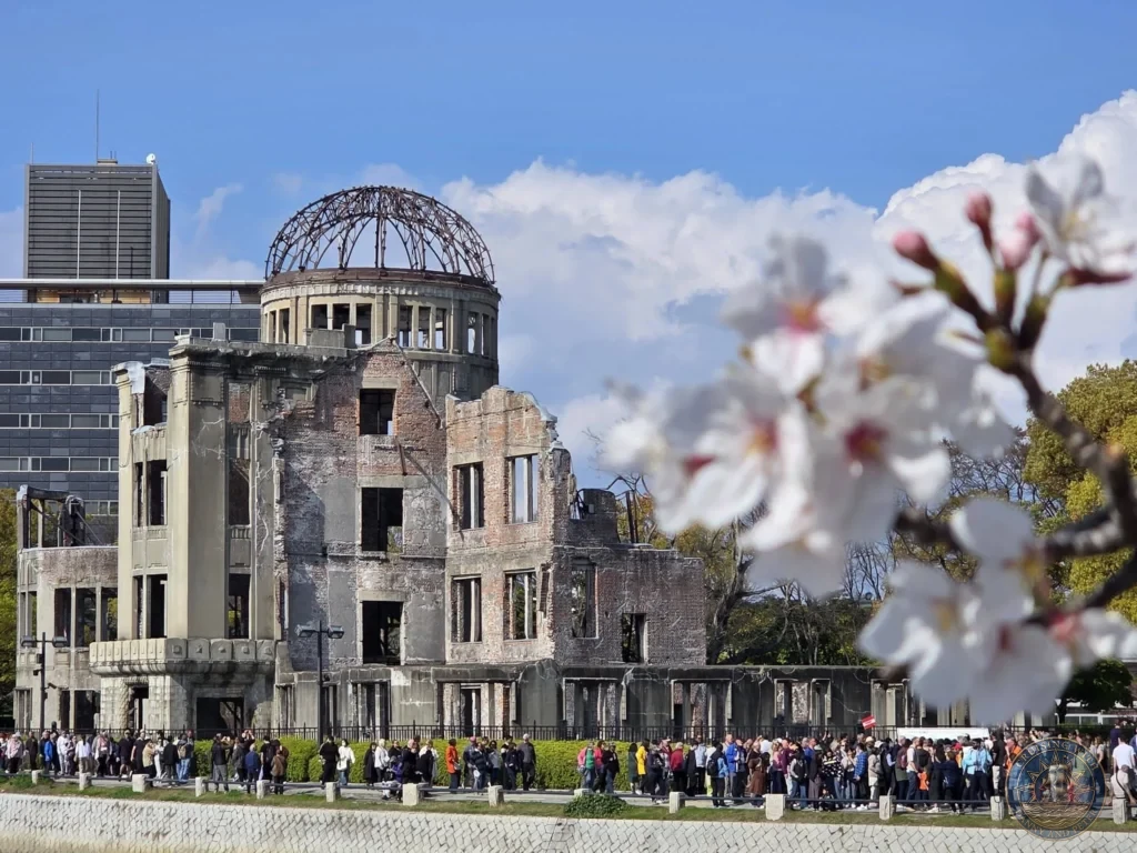 Hiroshima Japan Cruise Port Guide 2026 – Insider Tips & One Day Itinerary 8 Soft white cherry blossoms blooming in the foreground framing the Atomic Bomb Dome and river promenade crowded with visitors in Hiroshima Japan. Hiroshima Japan Cruise Port Guide, DIY Hiroshima Cruise Port to City Center, Things to do in Hiroshima from cruise ship, Westerdam Hiroshima Docking Location, One day in Hiroshima Japan itinerary by Danny and Peter.