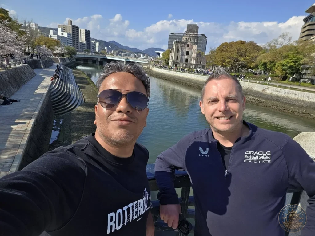 Hiroshima Japan Cruise Port Guide 2026 – Insider Tips & One Day Itinerary 51 Selfie of Danny and Peter smiling on a bridge over the Motoyasu River with the Atomic Bomb Dome and cherry blossom trees visible in the background in Hiroshima Japan. Hiroshima Japan Cruise Port Guide, DIY Hiroshima Cruise Port to City Center, Things to do in Hiroshima from cruise ship, Westerdam Hiroshima Docking Location, One day in Hiroshima Japan itinerary by Danny and Peter.