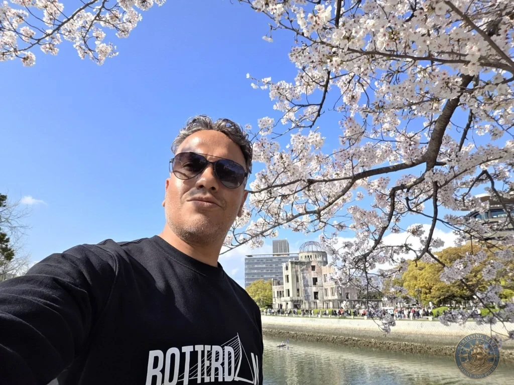 Hiroshima Japan Cruise Port Guide 2026 – Insider Tips & One Day Itinerary 52 Selfie of Danny framed by blooming cherry blossom branches with the Atomic Bomb Dome across the Motoyasu River in Hiroshima Japan. Hiroshima Japan Cruise Port Guide, DIY Hiroshima Cruise Port to City Center, Things to do in Hiroshima from cruise ship, Westerdam Hiroshima Docking Location, One day in Hiroshima Japan itinerary by Danny and Peter.