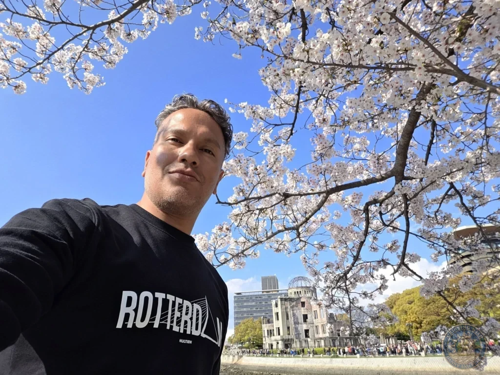 Hiroshima Japan Cruise Port Guide 2026 – Insider Tips & One Day Itinerary 53 Selfie of Danny framed by a full canopy of blooming cherry blossom branches with the Atomic Bomb Dome across the river on a clear day in Hiroshima Japan. Hiroshima Japan Cruise Port Guide, DIY Hiroshima Cruise Port to City Center, Things to do in Hiroshima from cruise ship, Westerdam Hiroshima Docking Location, One day in Hiroshima Japan itinerary by Danny and Peter.