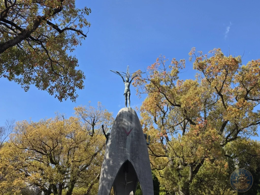 Hiroshima Japan Cruise Port Guide 2026 – Insider Tips & One Day Itinerary 55 Low angle view of the Children's Peace Monument statue holding a wire crane against a bright blue sky surrounded by trees in Hiroshima Peace Memorial Park Japan. Hiroshima Japan Cruise Port Guide, DIY Hiroshima Cruise Port to City Center, Things to do in Hiroshima from cruise ship, Westerdam Hiroshima Docking Location, One day in Hiroshima Japan itinerary by Danny and Peter.