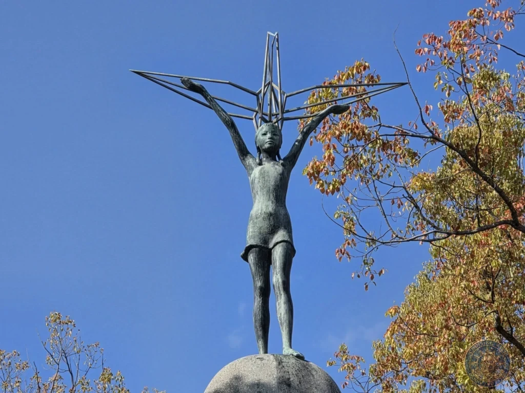 Hiroshima Japan Cruise Port Guide 2026 – Insider Tips & One Day Itinerary 56 Close-up of the bronze statue of a young girl holding a wire origami crane aloft against a clear blue sky at the Children's Peace Monument in Hiroshima Peace Memorial Park Japan. Hiroshima Japan Cruise Port Guide, DIY Hiroshima Cruise Port to City Center, Things to do in Hiroshima from cruise ship, Westerdam Hiroshima Docking Location, One day in Hiroshima Japan itinerary by Danny and Peter.