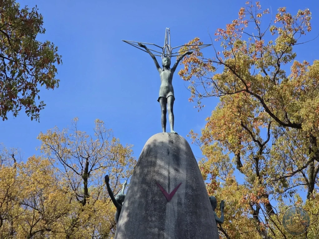 Hiroshima Japan Cruise Port Guide 2026 – Insider Tips & One Day Itinerary 57 View of the bronze statue of Sadako atop the curved pedestal with side figures framed by colorful trees against a clear blue sky at the Children's Peace Monument in Hiroshima Peace Memorial Park Japan. Hiroshima Japan Cruise Port Guide, DIY Hiroshima Cruise Port to City Center, Things to do in Hiroshima from cruise ship, Westerdam Hiroshima Docking Location, One day in Hiroshima Japan itinerary by Danny and Peter.