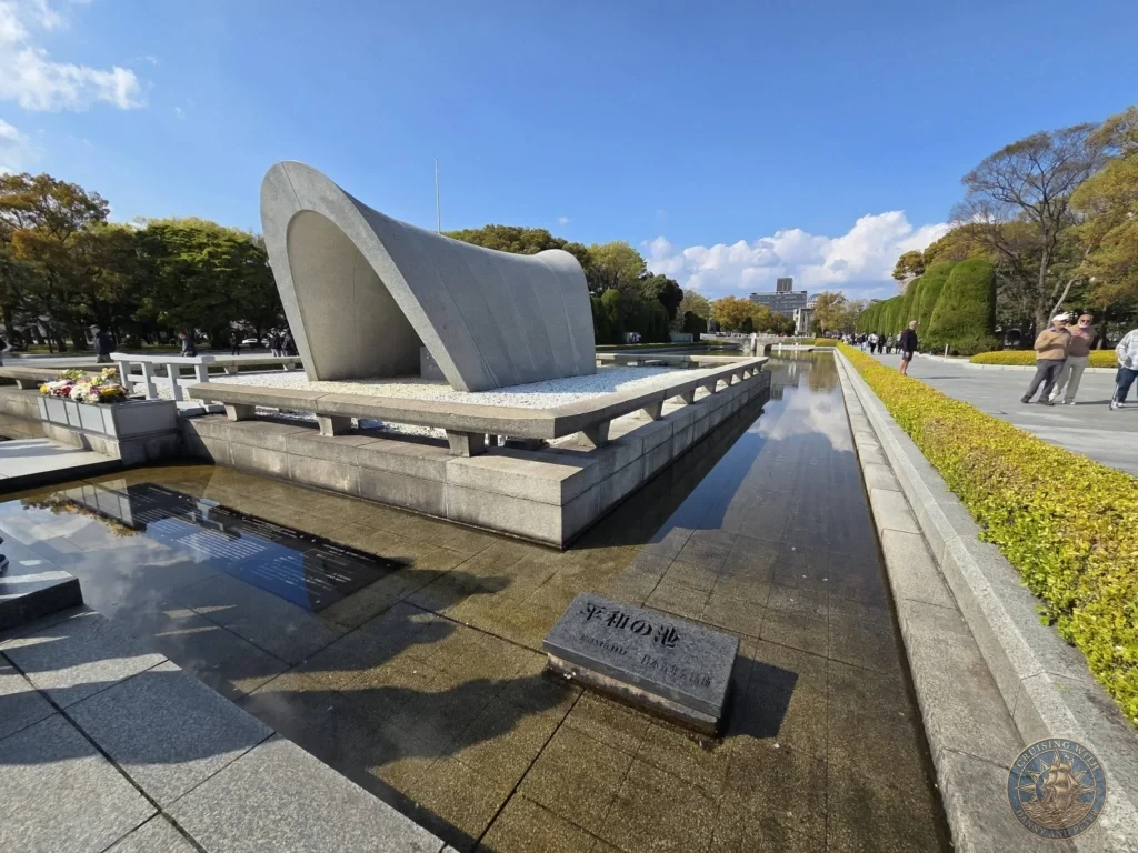 Hiroshima Japan Cruise Port Guide 2026 – Insider Tips & One Day Itinerary 17 Wide angle view of the arched Flame of Peace monument sitting in the Pond of Peace with surrounding walkways under a blue sky at Hiroshima Peace Memorial Park Japan. Hiroshima Japan Cruise Port Guide, DIY Hiroshima Cruise Port to City Center, Things to do in Hiroshima from cruise ship, Westerdam Hiroshima Docking Location, One day in Hiroshima Japan itinerary by Danny and Peter.