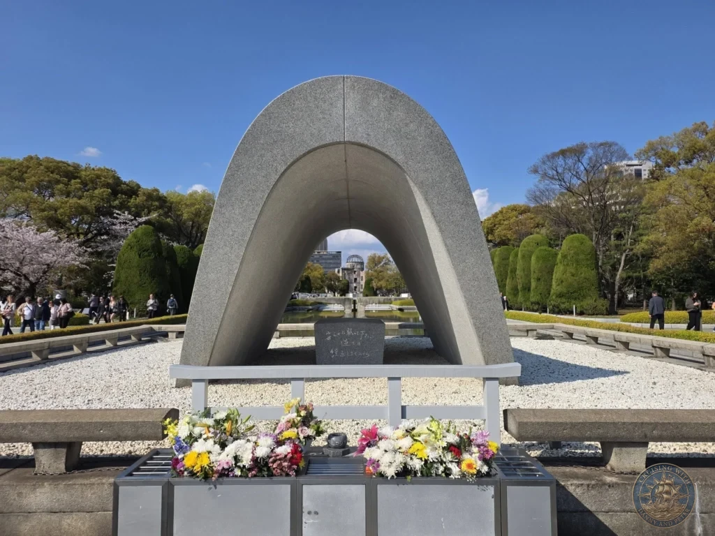 Hiroshima Japan Cruise Port Guide 2026 – Insider Tips & One Day Itinerary 58 Frontal view of the concrete arched Cenotaph for the A-Bomb Victims with flower offerings in the foreground, framing the Flame of Peace and the distant Atomic Bomb Dome in Hiroshima Peace Memorial Park Japan. Hiroshima Japan Cruise Port Guide, DIY Hiroshima Cruise Port to City Center, Things to do in Hiroshima from cruise ship, Westerdam Hiroshima Docking Location, One day in Hiroshima Japan itinerary by Danny and Peter.