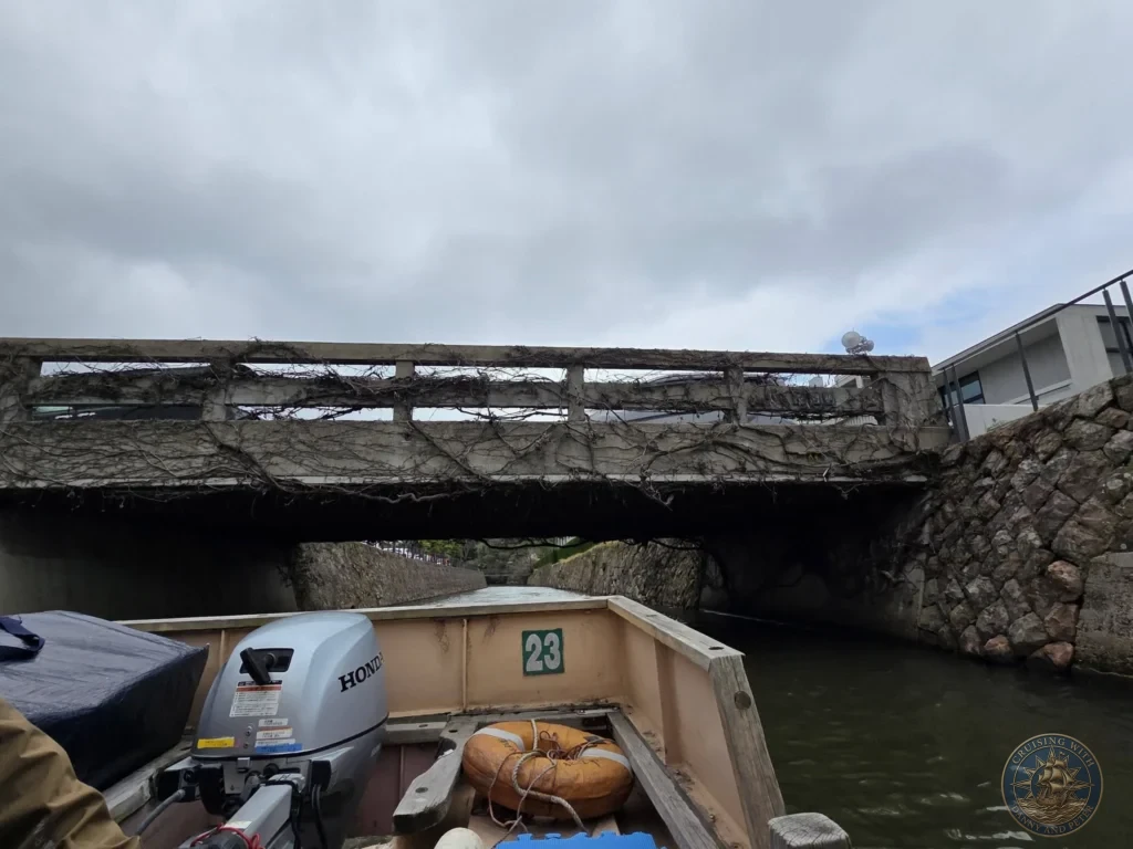 View from the stern of a Honda outboard motorboat passing under a low vine-covered concrete bridge along the Horikawa Canal in Matsue during a shore excursion from Sakaiminato Japan Cruise Port Guide, DIY Sakaiminato Cruise Port to City Center, Things to do in Sakaiminato from cruise ship, Westerdam Sakaiminato Docking Location, One day in Sakaiminato Japan itinerary by Danny and Peter.