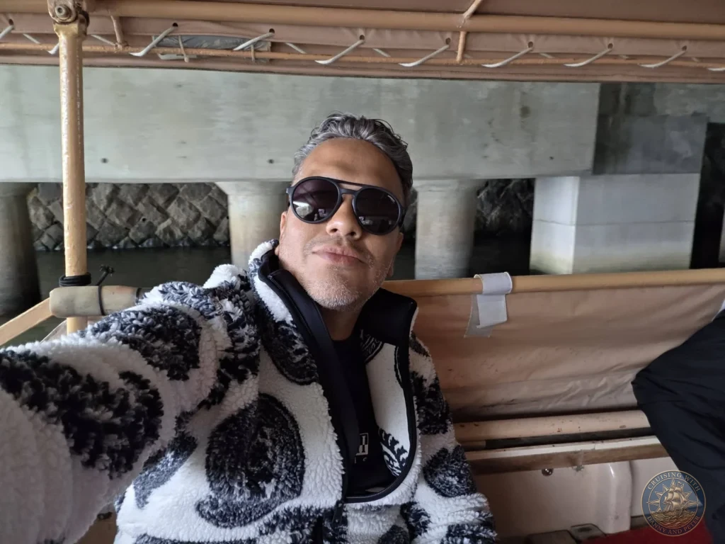 Danny wearing sunglasses and a patterned fleece jacket taking a selfie aboard the Horikawa Sightseeing Boat under a bridge in Matsue during a shore excursion from Sakaiminato Japan Cruise Port Guide, DIY Sakaiminato Cruise Port to City Center, Things to do in Sakaiminato from cruise ship, Westerdam Sakaiminato Docking Location, One day in Sakaiminato Japan itinerary by Danny and Peter.