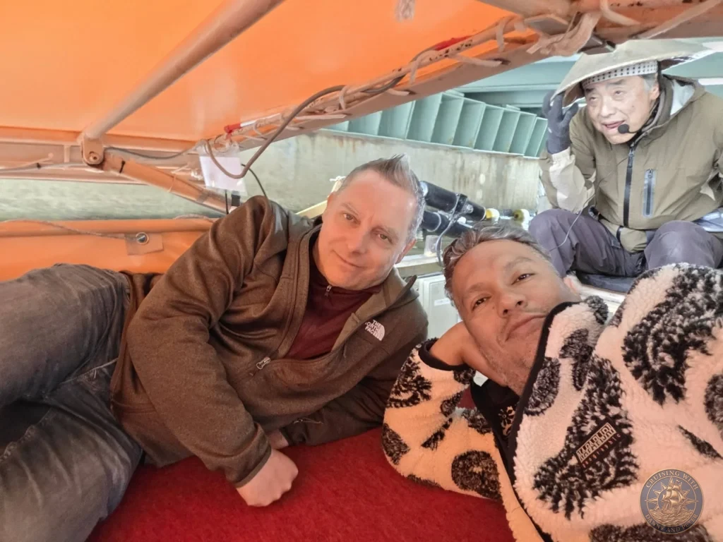 Danny and Peter lying flat inside the Horikawa Sightseeing Boat as the canopy lowers under a low bridge with the boat captain visible behind them in Matsue during a shore excursion from Sakaiminato Japan Cruise Port Guide, DIY Sakaiminato Cruise Port to City Center, Things to do in Sakaiminato from cruise ship, Westerdam Sakaiminato Docking Location, One day in Sakaiminato Japan itinerary by Danny and Peter.