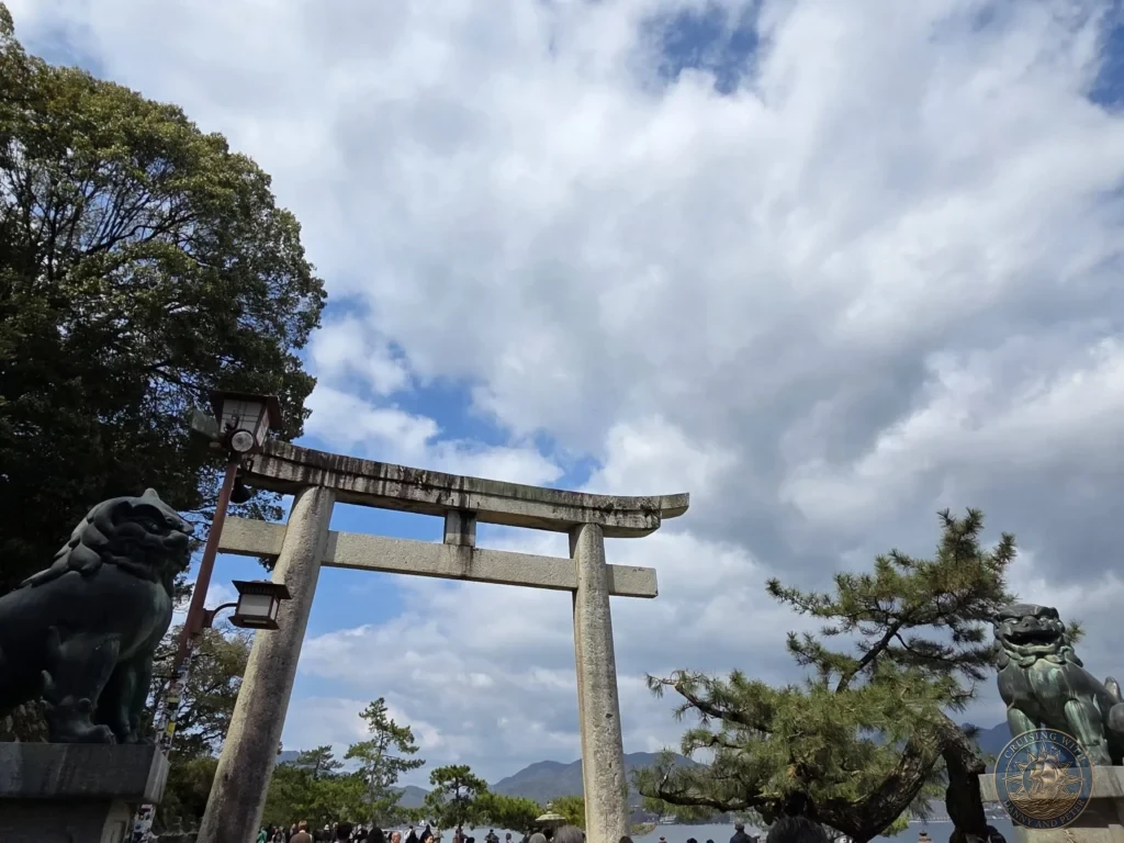 Hiroshima Japan Cruise Port Guide 2026 – Insider Tips & One Day Itinerary 36 Low-angle view of a large stone Torii gate flanked by guardian lion-dog statues under a dynamic cloudy sky on Miyajima Island in Hiroshima Japan. Hiroshima Japan Cruise Port Guide, DIY Hiroshima Cruise Port to City Center, Things to do in Hiroshima from cruise ship, Westerdam Hiroshima Docking Location, One day in Hiroshima Japan itinerary by Danny and Peter.