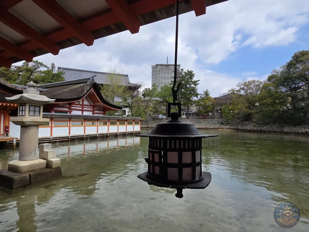 Hiroshima Japan Cruise Port Guide 2026 – Insider Tips & One Day Itinerary 79 Detailed view of a hanging bronze lantern and a stone lantern set over the water near the traditional wooden structures of Itsukushima Shrine on Miyajima Island in Hiroshima Japan. Hiroshima Japan Cruise Port Guide, DIY Hiroshima Cruise Port to City Center, Things to do in Hiroshima from cruise ship, Westerdam Hiroshima Docking Location, One day in Hiroshima Japan itinerary by Danny and Peter.