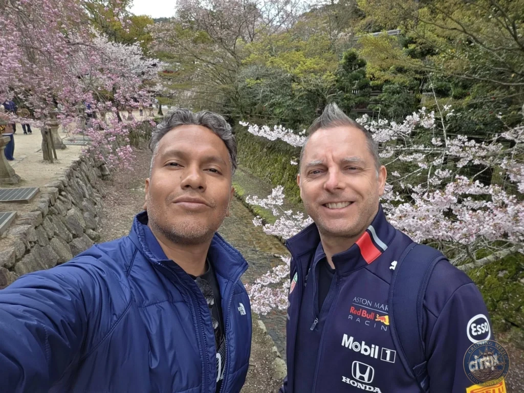 Hiroshima Japan Cruise Port Guide 2026 – Insider Tips & One Day Itinerary 77 Selfie of Danny and Peter smiling in front of blooming cherry blossom trees and a stone stream path on Miyajima Island in Hiroshima Japan. Hiroshima Japan Cruise Port Guide, DIY Hiroshima Cruise Port to City Center, Things to do in Hiroshima from cruise ship, Westerdam Hiroshima Docking Location, One day in Hiroshima Japan itinerary by Danny and Peter.