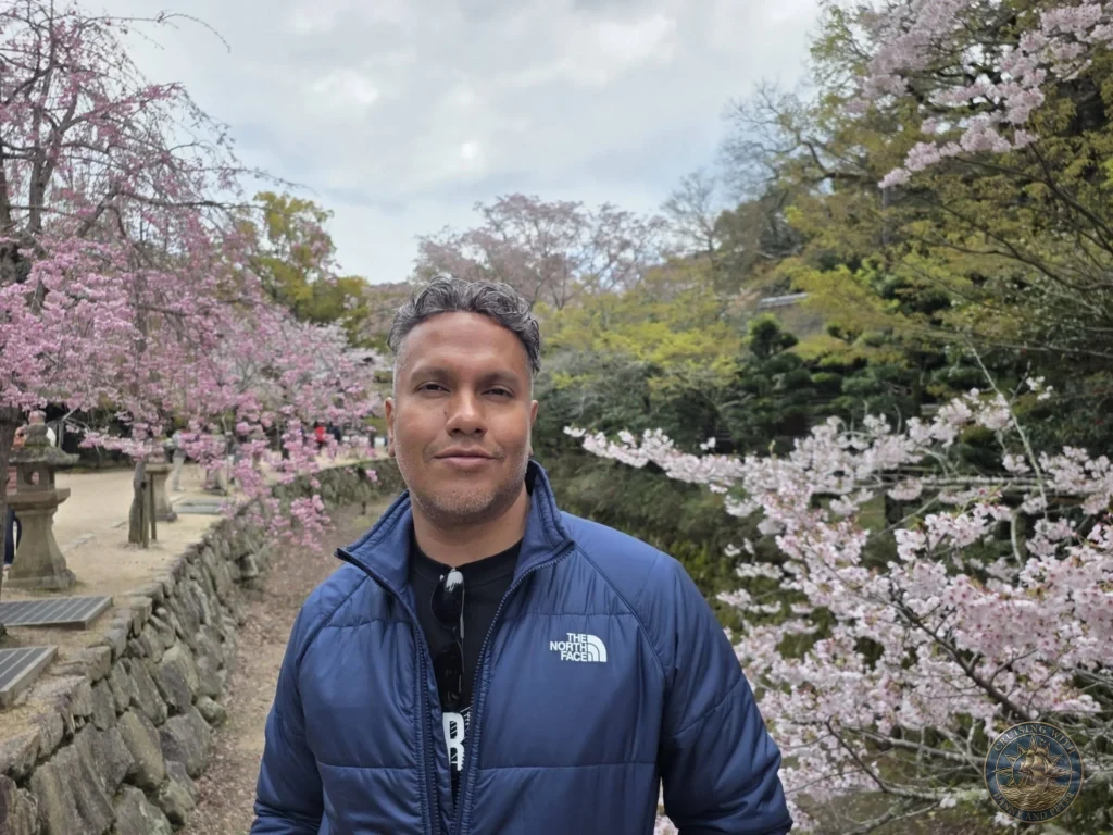 Hiroshima Japan Cruise Port Guide 2026 – Insider Tips & One Day Itinerary 43 Danny wearing a blue jacket standing in front of blooming pink cherry blossom trees and stone lanterns on Miyajima Island in Hiroshima Japan. Hiroshima Japan Cruise Port Guide, DIY Hiroshima Cruise Port to City Center, Things to do in Hiroshima from cruise ship, Westerdam Hiroshima Docking Location, One day in Hiroshima Japan itinerary by Danny and Peter.