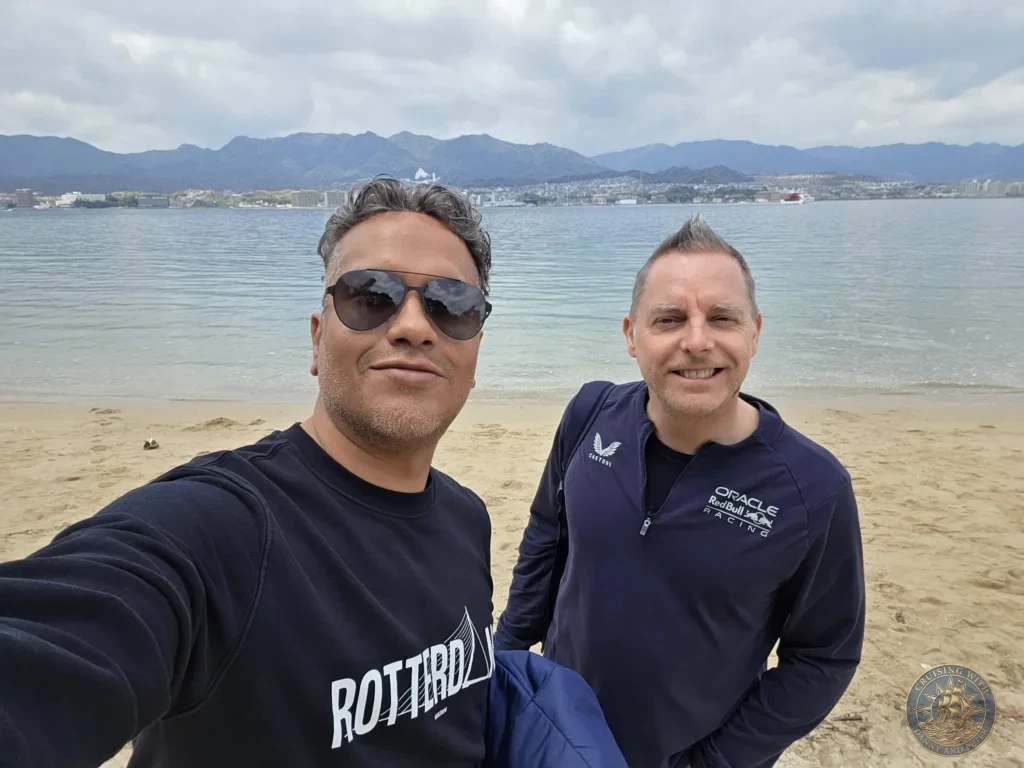 Hiroshima Japan Cruise Port Guide 2026 – Insider Tips & One Day Itinerary 46 Danny and Peter smiling on the sandy beach of Miyajima Island with Hiroshima Bay and distant mountains behind them in Hiroshima Japan. Hiroshima Japan Cruise Port Guide, DIY Hiroshima Cruise Port to City Center, Things to do in Hiroshima from cruise ship, Westerdam Hiroshima Docking Location, One day in Hiroshima Japan itinerary by Danny and Peter.