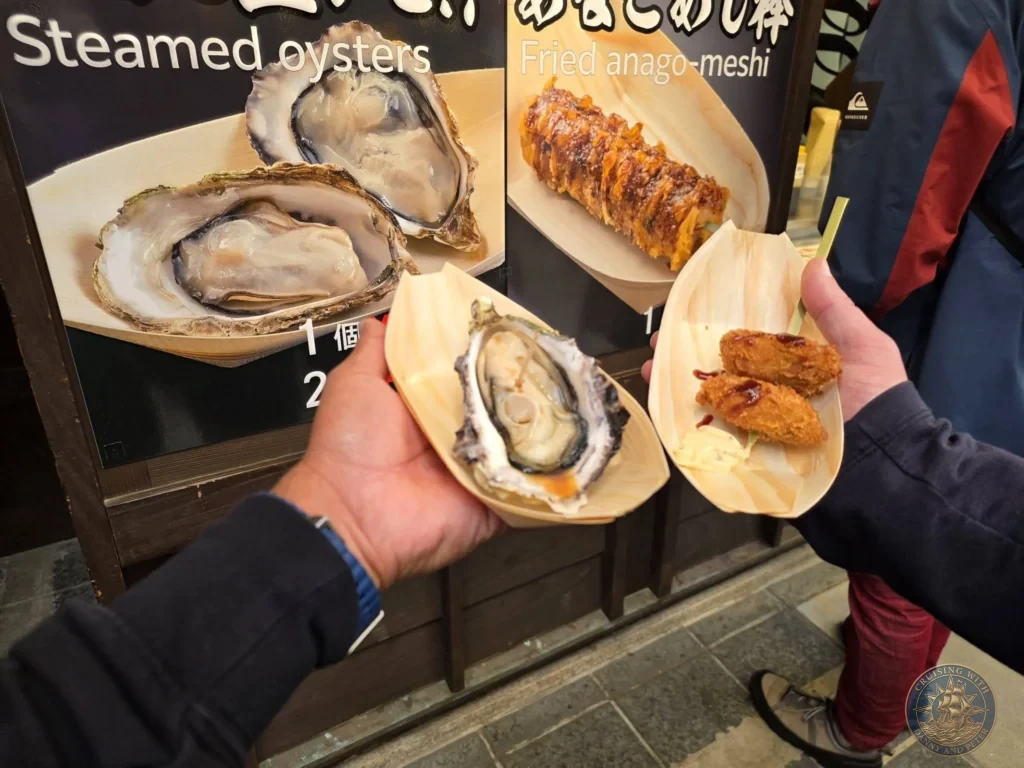 Hiroshima Japan Cruise Port Guide 2026 – Insider Tips & One Day Itinerary 19 Danny and Peter holding fresh steamed oysters and fried local seafood in wooden boat dishes at a street food stall on Miyajima Island in Hiroshima Japan. Hiroshima Japan Cruise Port Guide, DIY Hiroshima Cruise Port to City Center, Things to do in Hiroshima from cruise ship, Westerdam Hiroshima Docking Location, One day in Hiroshima Japan itinerary by Danny and Peter.