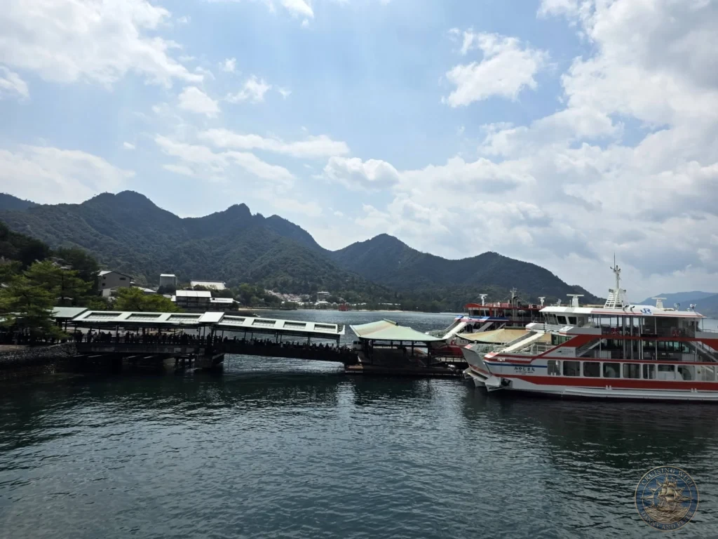Hiroshima Japan Cruise Port Guide 2026 – Insider Tips & One Day Itinerary 48 Ferries docked at the Miyajima Island pier terminal with covered boardwalk walkways, forested mountains, and a distant views of the floating torii gate in Hiroshima Japan. Hiroshima Japan Cruise Port Guide, DIY Hiroshima Cruise Port to City Center, Things to do in Hiroshima from cruise ship, Westerdam Hiroshima Docking Location, One day in Hiroshima Japan itinerary by Danny and Peter.